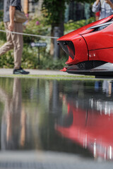 Sleek glossy red front end of sports car with stylish black mesh grille reflect in shallow pool at outdoor auto exhibition with visitors visible in bokeh background.