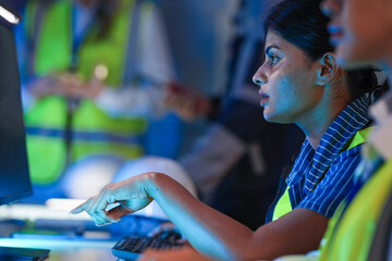 A stressed Indian female engineer in a safety vest works at a computer. She looks intently at the...