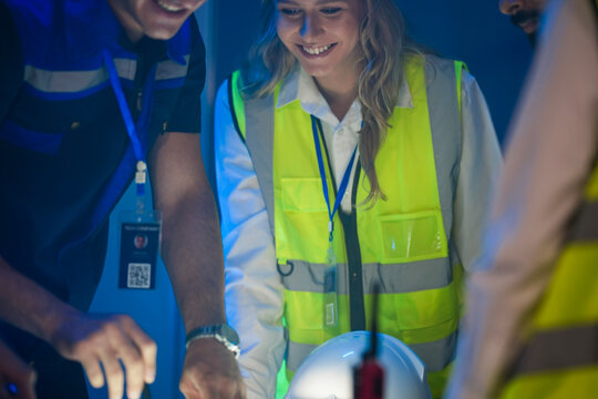 A happy, diverse team of engineers in safety vests collaborates at night. A smiling Caucasian woman and her colleagues look at a glowing blueprint in a factory control room. - Powered by Adobe