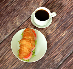 Cup of black coffee on saucer and sandwich of croissant with sausage, tomatoes and lettuce on wooden table.