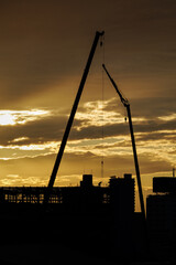 Two silhouetted crane arms frame warm sunset as lone rooftop worker receives parts hanging from crane's long suspension cable during site cleanup at end of another workday.