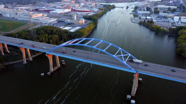 Traffic crosses the arched Leo Frigo Memorial Bridge as it stretches above the calm river lined with industrial buildings and autumn foliage in warm evening light.