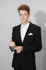 Young man in a black suit adjusting his jacket in a studio. Gray background.