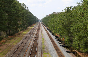 A set of railway tracks stretches straight ahead through a dense forest of green pine trees on both sides under a partly cloudy sky with some wooden beams on the ground