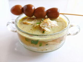 Indonesian chicken porridge (bubur ayam) served in a transparent glass bowl. The creamy, savory rice porridge is topped with shredded chicken and quail skewer,sliced green onions, and fried shallots.