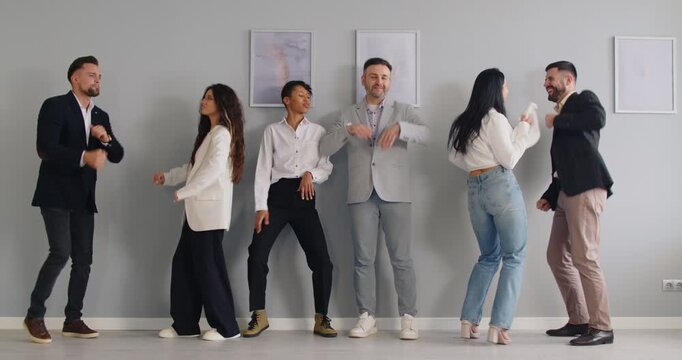Happy business people dance to music together at office wall. Team of young crazy employees dancing on party, cheerful corporate managers perform modern dance movements and fun jumps on meeting