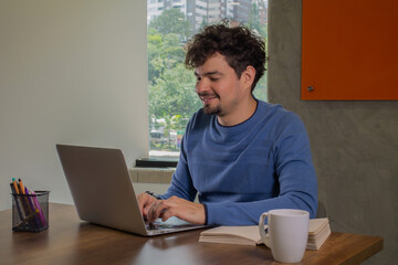 A portrait of a confident and smiling Latin man with curly hair sitting at a desk, enjoying a cup of coffee while working on his laptop in a creative office setting