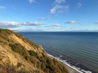 Pacific Ocean coast photo. High cliff with grass, shrubs. Sea of ​​Okhotsk with waves, ripples on the blue water. Summer landscape, sky with clouds. Sandy beach, coastline. For travel, tourism project