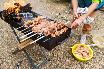 Person grilling meat skewers on a charcoal grill outdoors. Smoke and fire rising as juicy kebabs cook on skewers, summer picnic cooking background.