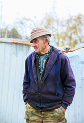 Elderly Russian man standing in a rural village, hands in pockets, looking sideways, dressed in casual clothes and hat.