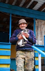 Elderly Russian man standing on the porch of his rural home, holding his gray and white cat with care and warmth, simple village life moment.