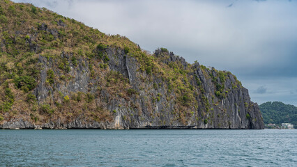 Rocky coast of a tropical island. Green vegetation grows on the steep slopes of high cliffs. Clouds in the sky. Buildings on the shore in the distance. Philippines. Palawan. El Nido