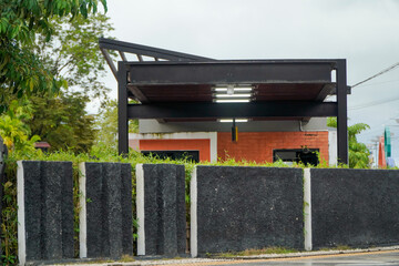 A modern building with a dark metal roof structure and orange accents, partially obscured by a textured black and white concrete wall and lush green foliage.