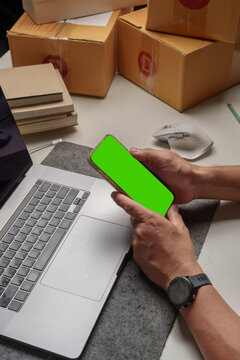 Close-up of a person holding a smartphone with a green screen on a work desk beside a laptop and delivery boxes, suitable for e-commerce, online business, and mockup design.