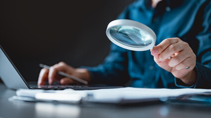 A business professional using a magnifying glass to review and verify documents on a desk,...