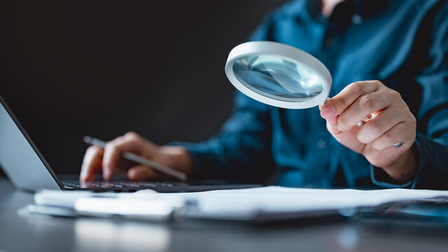 A business professional using a magnifying glass to review and verify documents on a desk, symbolizing audit, compliance, data verification, and quality assurance.
