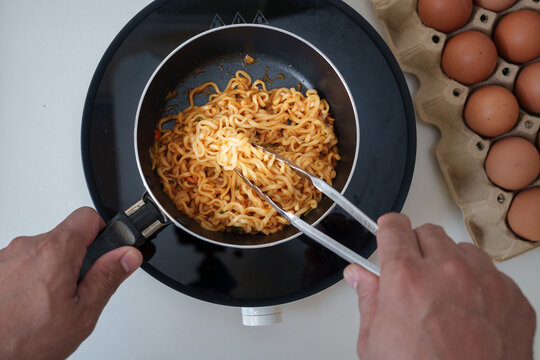 Top view of a person cooking instant noodles in a pot using tongs with eggs beside, representing simple homemade food, quick meal preparation, and Asian cuisine concept.