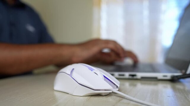 White Computer Mouse and Laptop in Office Setting, Close-up of a white computer mouse on a desk with a person typing on a laptop in the background