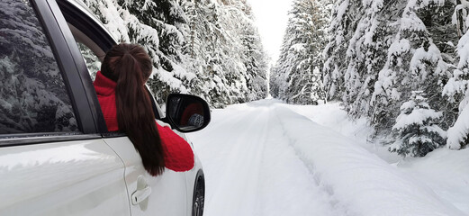 Young woman with red sweater enjoying a road trip in the white car on a snowy forest. Space for text. Wintertime, coming home for Christmas, enjoying, relaxing, tourism, travel, leisure time concept. 