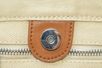 detail of a silver magnetic snap button on a brown leather patch, stitched onto cream canvas with a metal zipper below. Represents fashion hardware, textile craft, and bag accessories.