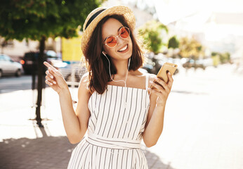 Portrait of happy smiling teen in summer hipster clothes listening music on her headphones from smartphone in the street on a sunny day