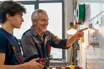 Experienced senior electrician mentor teaching a young apprentice about electrical wiring diagrams and circuits in a workshop, pointing to the board