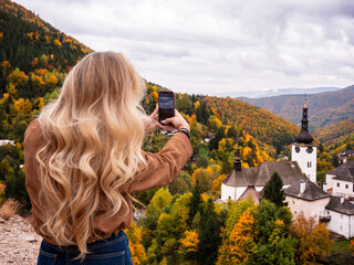 Young woman with long wavy blonde hair taking photo of Špania Dolina village in Slovakia during autumn. Stylish look, natural beauty and scenic mountain landscape. © Marek
