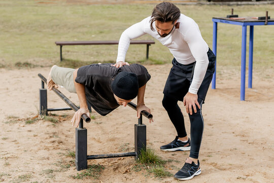 Two young men exercising outdoors on parallel bars in a park, one performing a push-up on the bars while the other watches and encourages, showcasing friendship, fitness, and outdoor training
