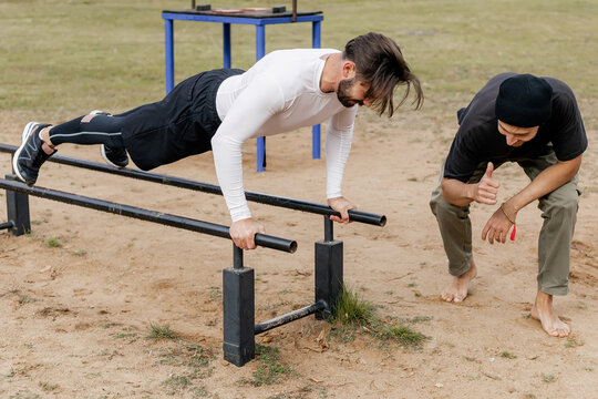 Two young men exercising outdoors on parallel bars in a park, one performing a push-up on the bars while the other watches and encourages, showcasing friendship, fitness, and outdoor training