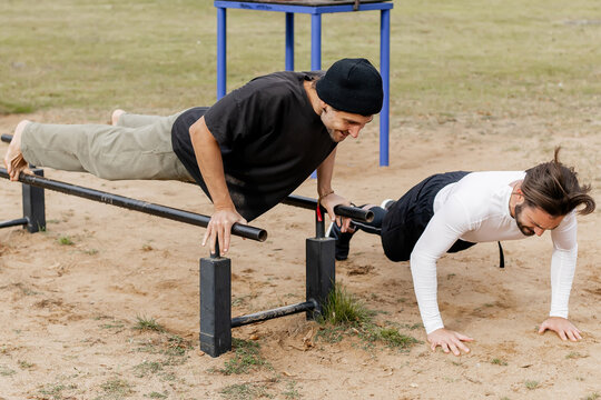 Two young men exercising outdoors on parallel bars in a park, one performing a push-up on the bars while the other watches and encourages, showcasing friendship, fitness, and outdoor training