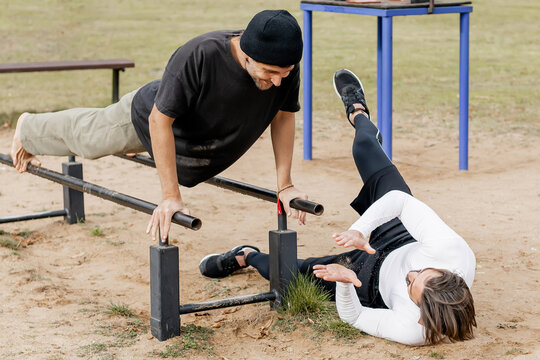 Two young men exercising outdoors on parallel bars in a park, one performing a push-up on the bars while the other watches and encourages, showcasing friendship, fitness, and outdoor training