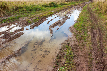 Muddy rural dirt road with deep ruts and reflective puddles under a cloudy sky, bordered by autumn grass