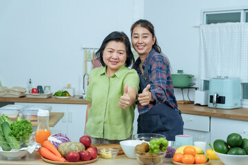 Senior asian mother adult daughter smiling happily showing thumbs up in kitchen after preparing healthy salad surrounded by colorful fruits vegetables representing success love and family connection