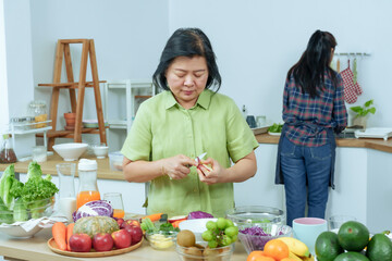 Asian senior mother peeling fruit at kitchen table with fresh vegetables while adult daughter working behind symbolizing healthy lifestyle family teamwork love and domestic happiness bonding together