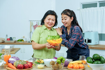 Asian senior mother showing salad bowl proudly while adult daughter smiling beside enjoying cheerful family moment representing love happiness bonding health wellbeing and positive lifestyle at home