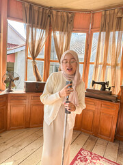 A young woman is singing into a vintage microphone in a bright, charming room with large windows