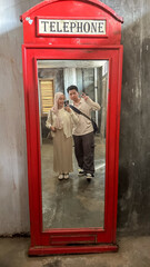 A couple stands side by side, smiling and posing for a reflection in a vintage red booth mirror