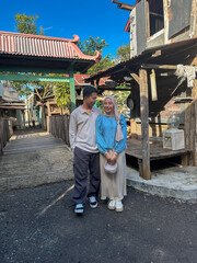 A couple stands side by side in a charming village, smiling under the bright blue sky