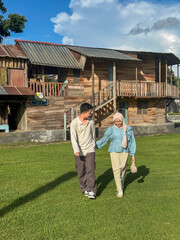 Two people walk hand in hand on a bright day near a wooden house, surrounded by green grass