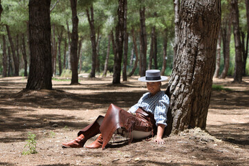 A young, pretty woman dressed in riding breeches and riding clothes and a typical Spanish hat rests seated in the shade of a pine tree on a pilgrimage day in Spain.
