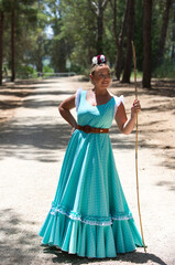 Fototapeta premium A beautiful young Spanish woman dressed in a traditional green flamenco dress and with a flower in her hair walks through the pine forests of Andalusia in the pilgrimage, leaning on a stick.