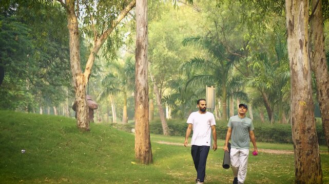 Two handsome men carrying a mattress walk along a park trail, talking to each other amid green grass and rows of trees. A joyful moment captured in slow motion at Delhi, India.
