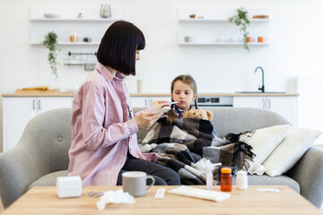 A concerned mother checks her daughter's temperature while she is wrapped in a blanket on the sofa, showing care and concern.