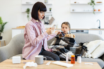 A concerned mother checks her daughter's temperature while she is wrapped in a blanket on the sofa, with medicine on the table.