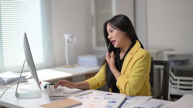 A woman in a yellow jacket is talking on her cell phone while sitting at a desk. She is typing on a computer keyboard and has a stack of papers in front of her. Concept of productivity and focus