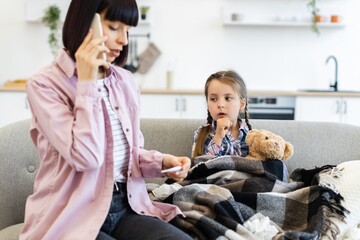 A concerned mother talks on the phone while checking her daughter's temperature, who is wrapped in a blanket on the couch, looking unwell.