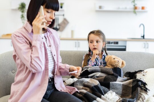 A concerned mother talks on the phone while checking her daughter's temperature, who is wrapped in a blanket on the couch, looking unwell.