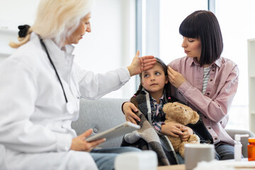 A doctor checks a young girl's temperature while her mother comforts her. The child is wrapped in a blanket and holding a teddy bear.