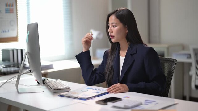 A woman is sitting at a desk with a computer monitor in front of her. She is wearing a suit and she is working on a project