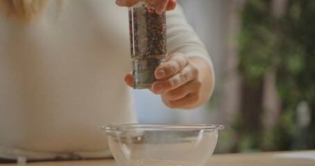 A person using a pepper grinder to add seasoning into a clear glass bowl, prepping ingredients for...